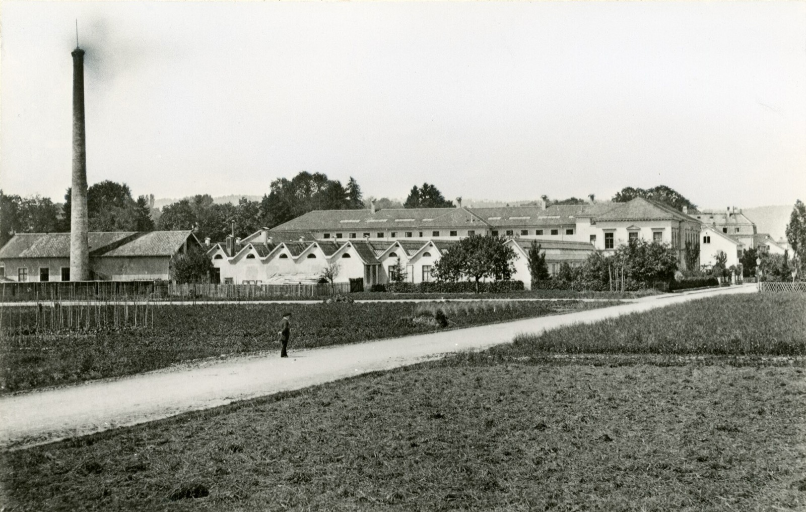 Historische Schwarz-Weiss-Fotografie der Seidenstoff-Weberei in Winterthur um 1880: langgezogene Fabrikgebäude mit Sheddächern und ein hoher rauchender Fabrikkamin, davor Felder und ein einzelner Mann auf einem Feldweg.