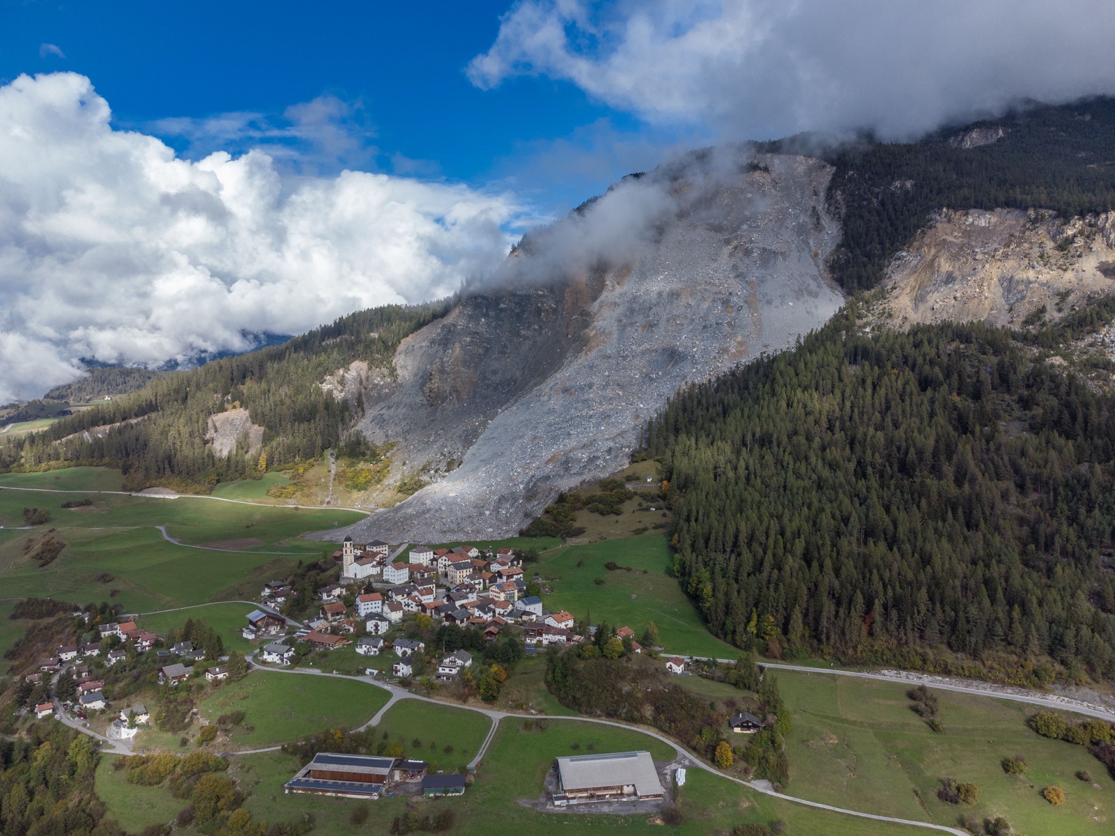 Luftaufnahme des Dorfes Brienz/Brinzauls in Graubünden: Eine breite graue Schutt- und Felszunge des Bergsturzes von 2023 reicht den Hang hinunter und endet unmittelbar vor den ersten Häusern des Dorfes.
